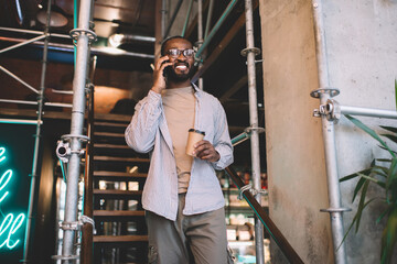 Joyful black hipster guy in optical eyeglasses smiling during friendly mobile talking on leisure, happy African American man making contact cell calling via smartphone application during coffee time