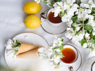 White tea set with tea in cups with yellow lemons and a blossoming apple branch.View from aboveThere are waffle cones on a saucer.