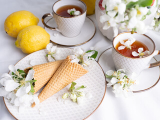 Obraz na płótnie Canvas White tea set with tea in cups with yellow lemons and a blossoming apple branch..There are waffle cones on a saucer.