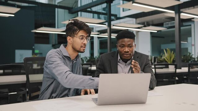 Two multicultural professional male co-workers men colleagues sit at office use laptop discuss internet project hold negotiation choosing business profitable contract cooperation teamwork partnership