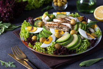 Salad with avocado, chicken, eggs and olives, dressed with olive oil, served on lettuce leaves on a dark gray background