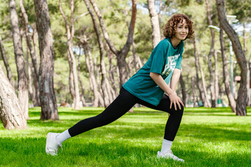 Full length shot of young redhead woman wearing green t-shirt and black legging standing on city park, outdoors doing stretching workout. Stretching and motivation, outdoor sport concepts.