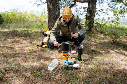 A Man On A Hike Prepares Delicious Food, Boil Water On A Gas Burner In A Pot, Camping Equipment, A Guy Cook Looks At The Readiness Of Food.