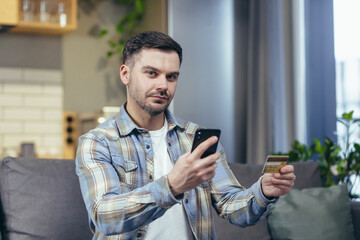 A man at home sitting on the couch uses the phone for online shopping, holding a bank credit card