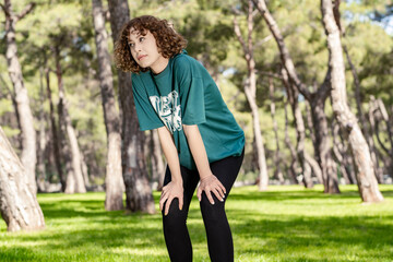 Young redhead woman wearing green t-shirt standing on city park, outdoor standing bent over and catching her breath after a running session. Outdoor sport concepts.