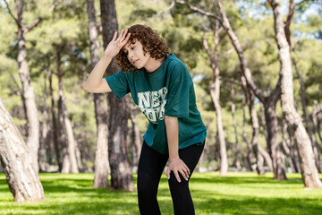 Young redhead woman wearing green t-shirt standing on city park, outdoor after jogging catching her breath, wiping her sweat from her forehead.