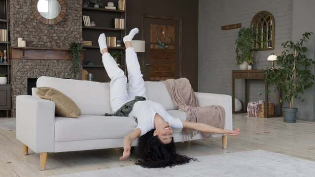 Young crazy funny carefree woman lying upside down on cozy sofa in living room shaking arms and legs dances lying enjoys being alone at home on weekend celebrating victory resting relaxing leisure