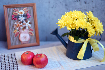 Still life of a bouquet of chrysanthemums in a decorative watering can, a picture in a frame and apples on a gray bokeh background. © Nadezda