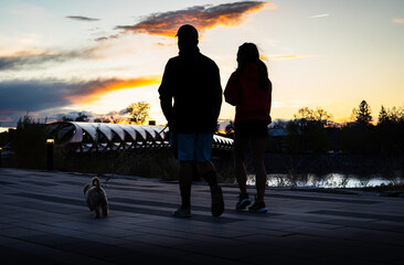 Calgary Alberta Canada, May 16 2022: A couple in love on a date night walking a puppy along a downtown bike path near the Peace Bridge with sunset lighting.