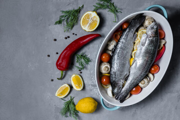 Raw trout in a blue baking dish, on a gray table surrounded by lemons, peppers, mushrooms and tomatoes, top view. Wild Fish, preparation for cooking. The concept of healthy eating. 