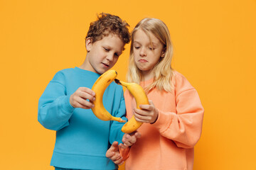 funny, cute children, a boy and a girl stand on a blue background in bright clothes and holding bananas in their hands are fooling around, connecting fruits in the shape of a circle