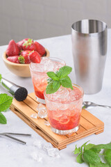 Cold summer strawberry cocktail mojito, margarita, daiquiri. Two glasses with fresh strawberry soda drink, ice cubes, mint and bartender tools on light background. Iced strawberry lemonade, close up