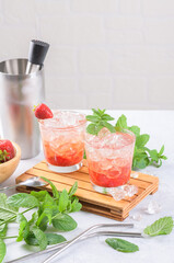 Cold summer strawberry cocktail mojito, margarita, daiquiri. Two glasses with fresh strawberry soda drink, ice cubes, mint and bartender tools on light background. Iced strawberry lemonade, close up