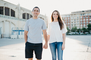 A Hispanic man and a smiling brunette are holding their hands near the pier in Spain in the evening. A couple of tourists on a date at the sunset in Valencia.
