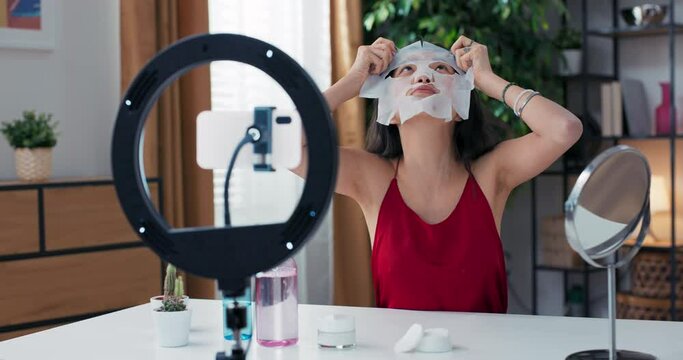 Beauty blogger is live streaming to review makeup products cosmetics on social media while applying fabric beauty mask to face. Influencer uses smartphone on a tripod and a ring light.