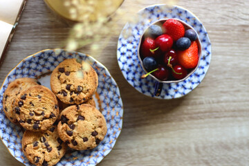 Plate of chocolate chip cookies, cup filled with strawberries, blueberries and cherries, open book and vase with gypsophila flowers on the table. Flat lay.