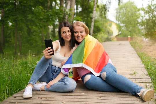 Two lesbian friends are photographed together in a park with an LGBT flag on their shoulder. June is the Month of pride and equality of human rights. Happy cute girls with a phone in  hand