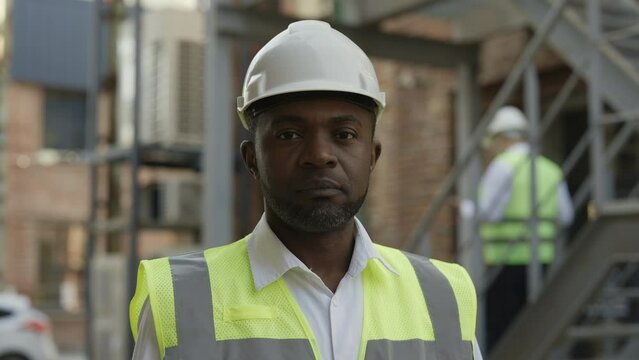 Portrait of african american male builder in hardhat looking at camera with calm face outdoor. Confident man working in large construction company.