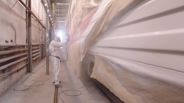 A man in a protective suit and a respirator covers the car with a protective layer of varnish using a pulverizer. Work at the carriage plant in the paint shop