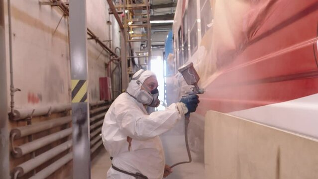 A man in a tattered old hazmat suit and respirator coats a train car with a protective layer of lacquer using a pulverizer. Toxic work at the car factory in the paint shop