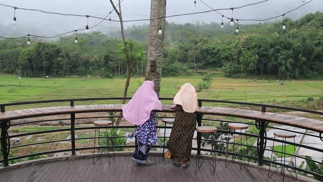 Two women wearing hijab while enjoying the resort atmosphere with views of rice fields and forests