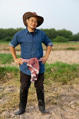 Fototapeta premium Handsome Asian man farmer is at plantation. He wears hat, blue shirt, Thai loincloth on waist, boots, put hands on hips, feels confident, looks at camera. Concept : Agriculture occupation. Full body.