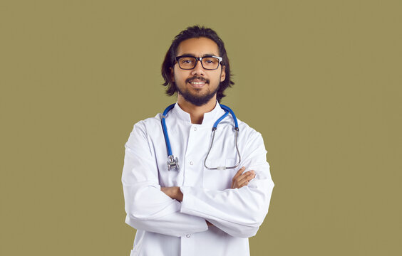 Portrait Of Male Doctor. Studio Shot Of Young Indian Physician. Confident Handsome South Asian Man In White Coat With Stethoscope Standing With Arms Folded Isolated On Solid Greenish Brown Background