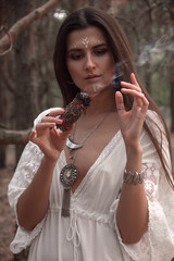 witch cosplay. close up young brunette girl in white boho dress and jewelry stands like modern witch with smoky burning stick in hand and makes shaman ritual on forest background. lifestyle concept