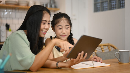 Smiling Asian mother helping her daughter doing homework, surfing internet on digital tablet at home
