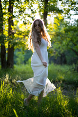 Beautiful young Russian girl dressed in a white dress, walks through the forest among the oaks in the rays of the sunset