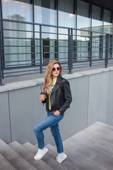 teen walk leisure. young blonde girl in jeans and black leather jacket stands in step on gray street stairs background with coffee in hand and looks apart in sunglasses. lifestyle concept, free space