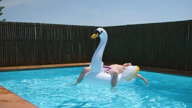Young happy boy relaxes and sunbathe on inflatable swan toy in blue pool.