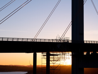 Fototapeta na wymiar Silhouette of suspension bridge and scaffolding for repair work. Shot into rising sun