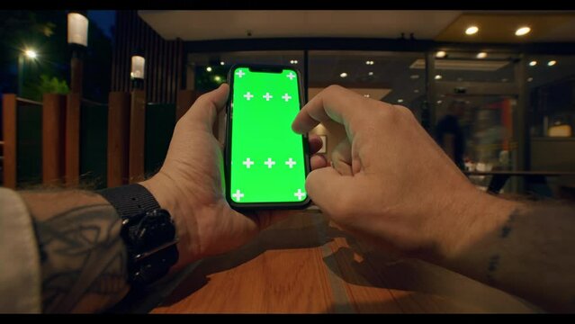 Close up front view of young male tattooed hands holding vertical and using mobile phone with green screen, swiping or scrolling smartphone touchscreen on table with soft lights of cafe on background