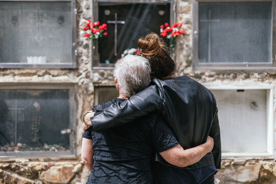 Elderly Widowed Mother And Orphaned Daughter Hugging To Comfort Each Other In Front Of The Grave At The Funeral Of The Husband And Father Of Both.concept Cemetery And Funeral Of Relative.are Crying.