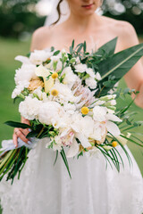 The bride in a white dress stands against a background of tropical plants and holds a bright bridal bouquet of flowers and greens 
