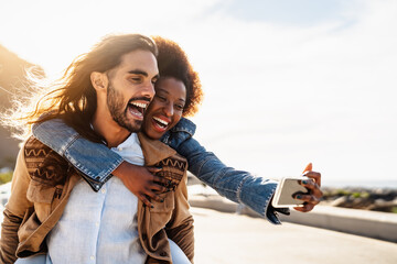 Happy multiracial couple having fun taking selfie with mobile smartphone during vacations - Youth...