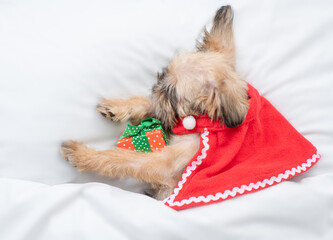 Brussels Griffon puppy wearing  red cape sleeps with gift box on a bed under white blanket at home....