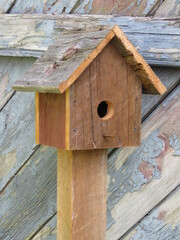 Simple Natural Wooden Birdhouse on Wood Post in Front of Old Weathered Gray Barn with Peeling Grey Paint