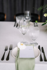 decoration of a wedding table, table setting for a wedding, white dishes on a wedding table with elements of green leaves and white flowers, themed decor. Orchid flower on a menu card