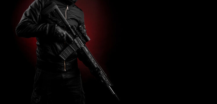 A man in dark clothes holds an automatic carbine in his hands. The face is covered with a mask, hands are in gloves. A figure with a weapon on a dark back with a red light spot.
