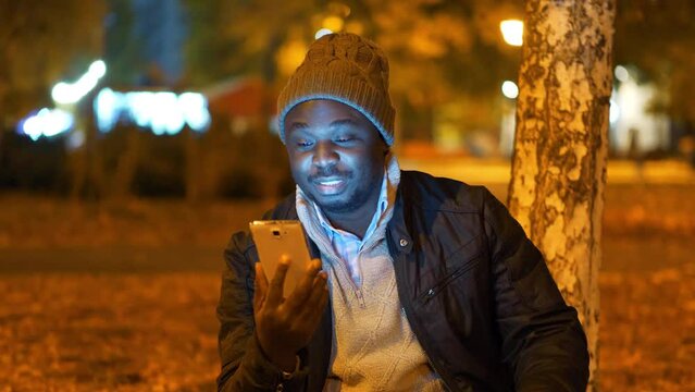 African american man in winter hat sits in evening city park and speaks via smartphone by video link. Video with flickering from outdoor lamps of artificial lighting