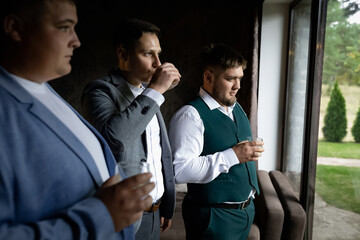 Three men with glasses of whiskey stand near the window in the apartment. Natural daylight. Groom's Gathering