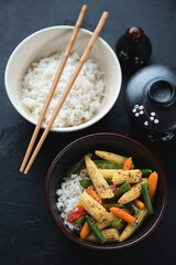 Bowls with panasian stir-fried vegetables and white rice, elevated view on a black stone background
