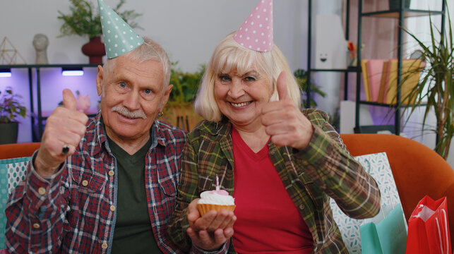 Happy senior family couple grandparents man woman wears festive birthday hat hold cupcake makes wish celebrating anniversary blowing burning candle on cake. Grandmother grandfather with gifts at home