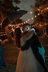 A couple in love under a black umbrella kisses. Curvy people, wedding couple on a walk. Stylish charming bride, groom in green suit. Beautiful park