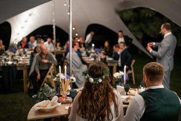 Wedding feast, large tent with guests. Fat people, wedding couple. Stylish charming bride, groom in green suit. Beautiful park