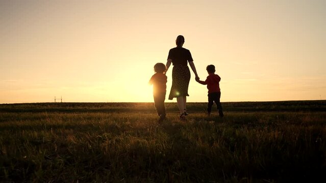 Silhouette of young family - mom and two brothers twins boys runs to the sun on open air field or park, golden hour, flares Happy mother and children, love, freedom, future concept