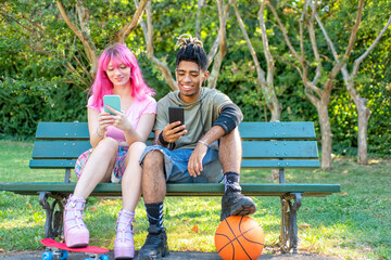 alternative diverse couple hanging out together sitting in a park bench holding smartphone online...