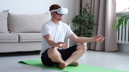 Young man using AR while meditating sitting in lotus position on roll mat wearing sports wear indoors. man wearing virtual reality device.